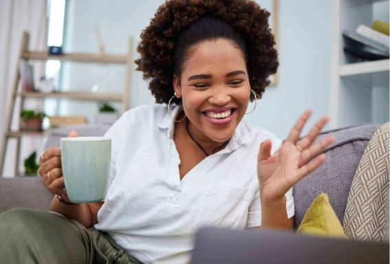 Total Mind - Psicólogos on-line e áudios de meditação Uma mulher em uma sala, sentada no sofá, sorrindo e segurando uma xícara, utilizando seu notebook à frente, como se estivesse em uma chamada de vídeo. #ParaTodosVerem