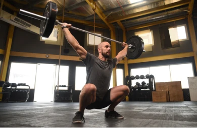 A foto está focada em um homem, que está dentro de um estúdio de cross training. Ele está fazendo agachamento com o auxílio de halter e pesos. #ParaTodosVerem