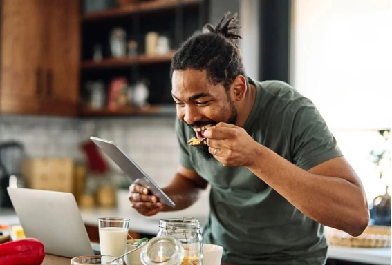 Total Nutri - Parceiros de Nutrição e conteúdos exclusivos Um homem na cozinha de casa, fazendo uma refeição, enquanto segura um tablet. Em frente, há uma mesa com alguns alimentos, como nozes, leite e frutas. #ParaTodosVerem